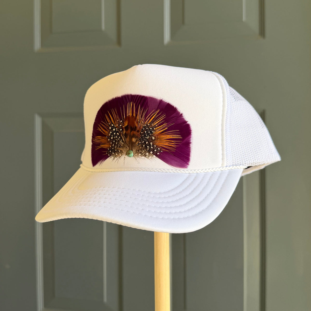 A white trucker-style cap displayed on a wooden stand in front of a gray paneled door. The front panel features a purple and gold feather fan design.