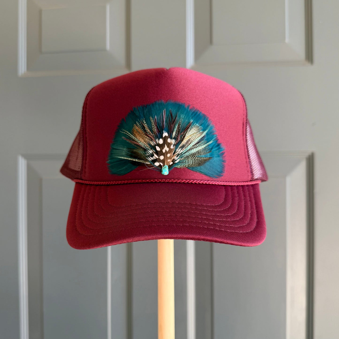 A burgundy maroon trucker hat with a teal and brown feather embellishment on the front, displayed on a wooden stand against a white paneled door.