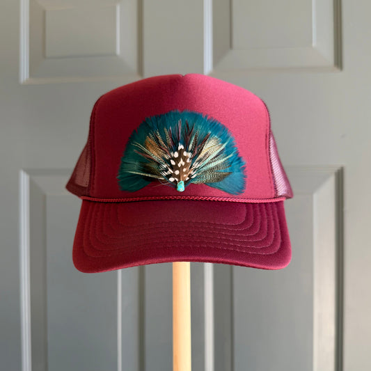 A burgundy maroon trucker hat with a teal and brown feather embellishment on the front, displayed on a wooden stand against a white paneled door.