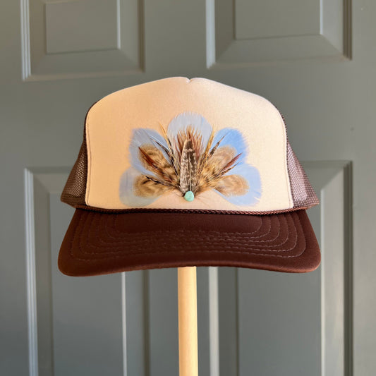 A trucker-style cap with a cream front panel, brown mesh sides, and a brown brim, decorated with a feathered flower design and a small turquoise bead on the front. The hat is displayed on a wooden stand against a gray paneled door.