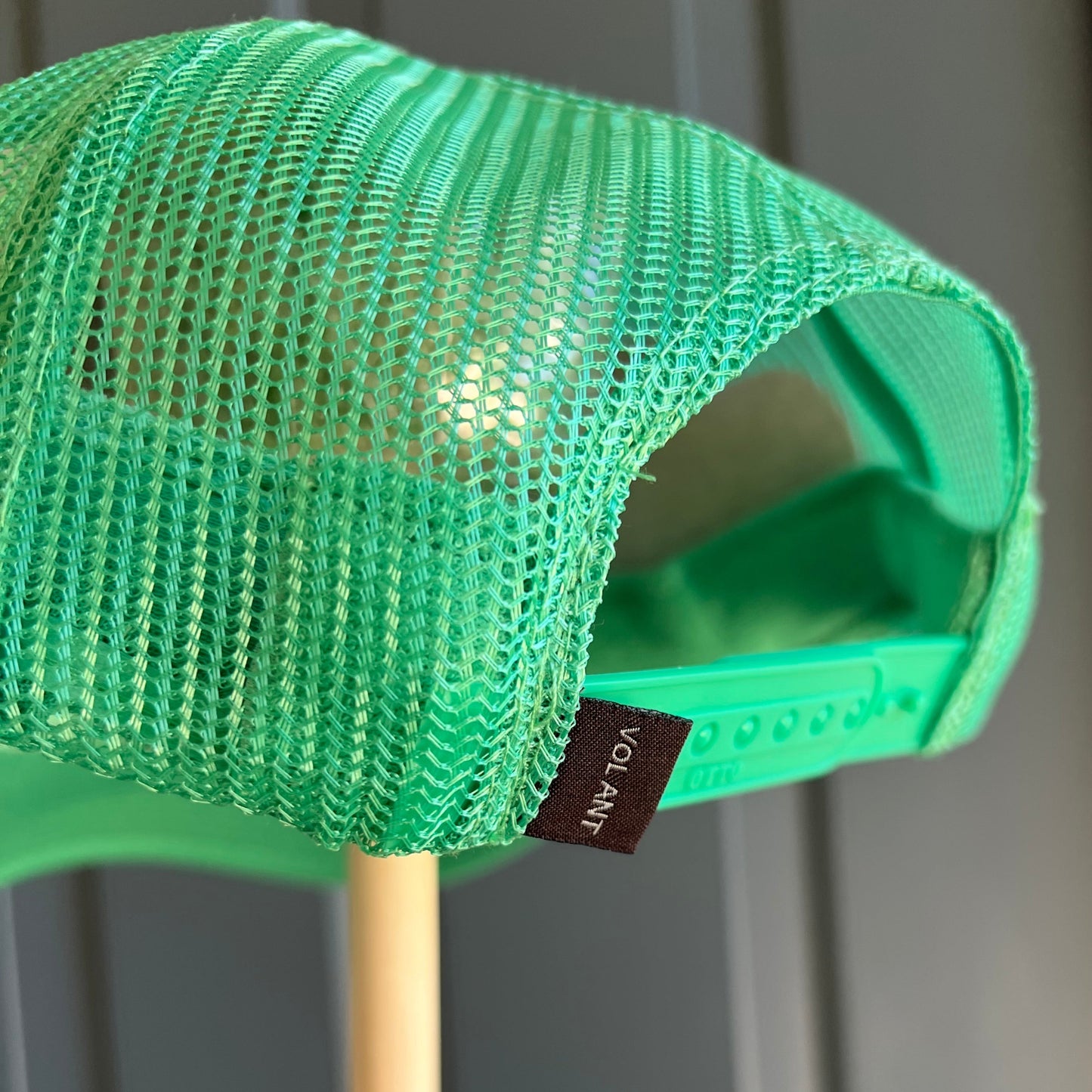 Close-up of a bright green mesh trucker hat showing the back panel, adjustable strap, and a small black brand tag reading VOLANT, with a light wood dowel in the background.