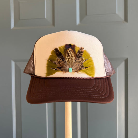 A brown and cream trucker hat with a decorative feather arrangement on the front panel, displayed on a wooden stand against a gray paneled door.