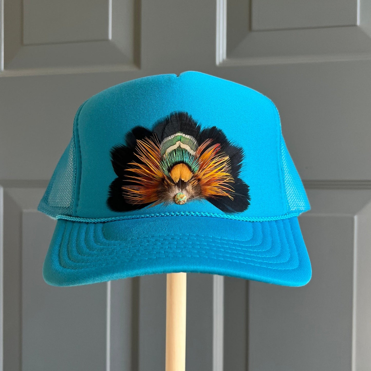 A bright turquoise blue trucker-style cap with a large decorative feather arrangement on the front, displayed on a wooden stand against a gray paneled door.