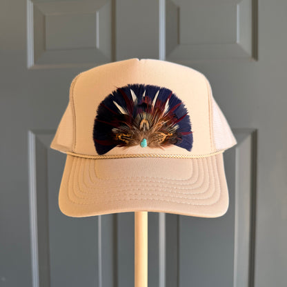 A beige trucker-style cap with a large decorative feather arrangement in navy, brown, and gold on the front, displayed on a wooden stand against a gray paneled door.
