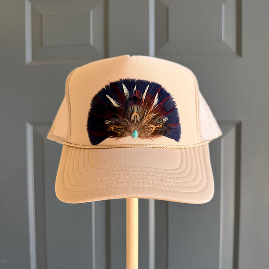 A beige trucker-style cap with a large decorative feather arrangement in navy, brown, and gold on the front, displayed on a wooden stand against a gray paneled door.