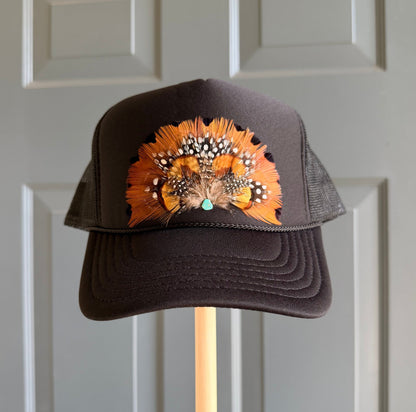 A black trucker hat with a vibrant orange and black feathered embellishment, displayed on a wooden stand against a light gray paneled door.