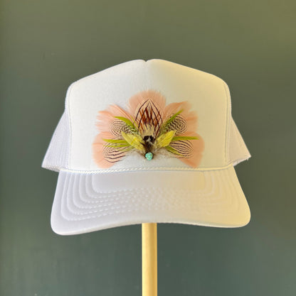 A white trucker-style cap displayed on a wooden stand against a dark green background, featuring a decorative pink and olive-green feather embellishment on the front.