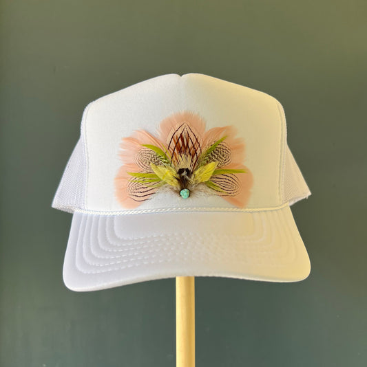 A white trucker-style cap displayed on a wooden stand against a dark green background, featuring a decorative pink and olive-green feather embellishment on the front.