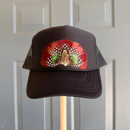 A black trucker hat with a curved brim and black mesh back panels, featuring a decorative feather patch on the front. The patch displays red, green, white, and gold feathers.