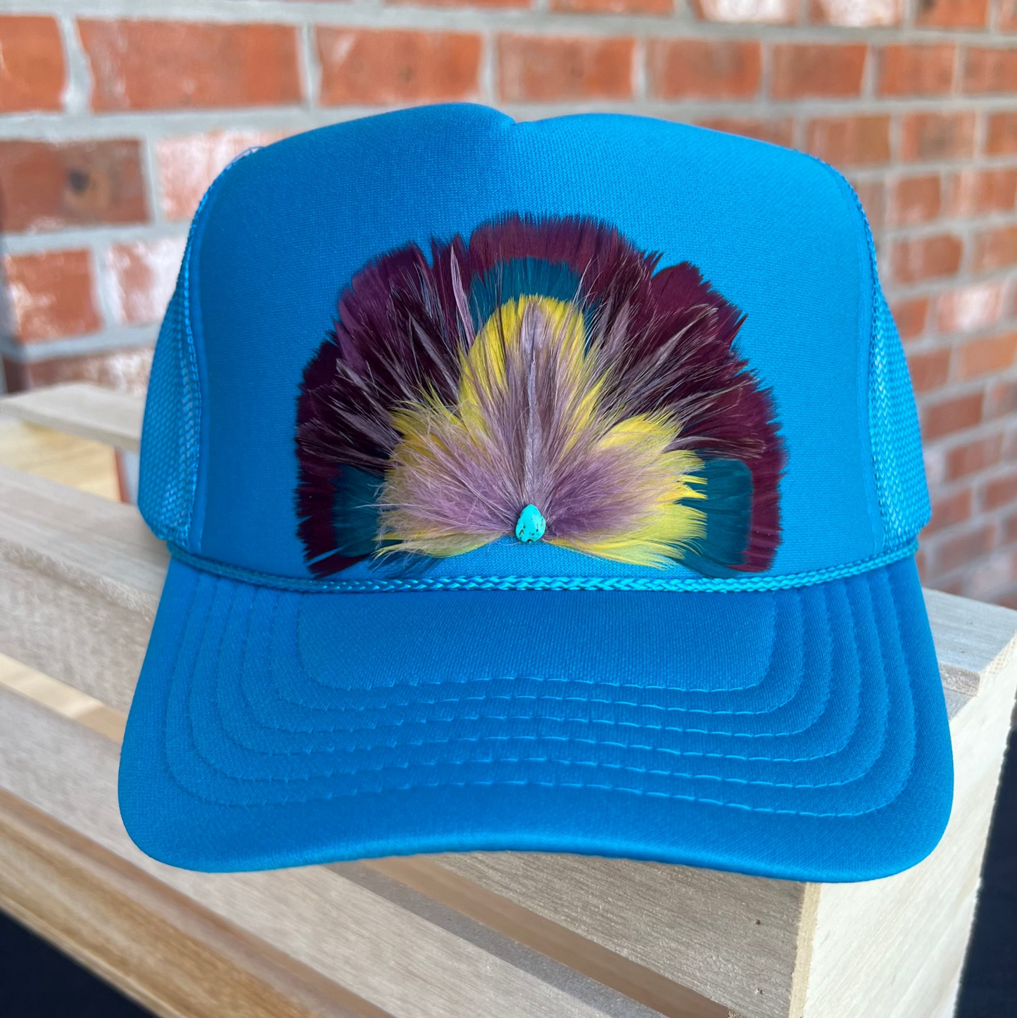 Blue cap with colorful feather design on a wooden surface with a brick wall background