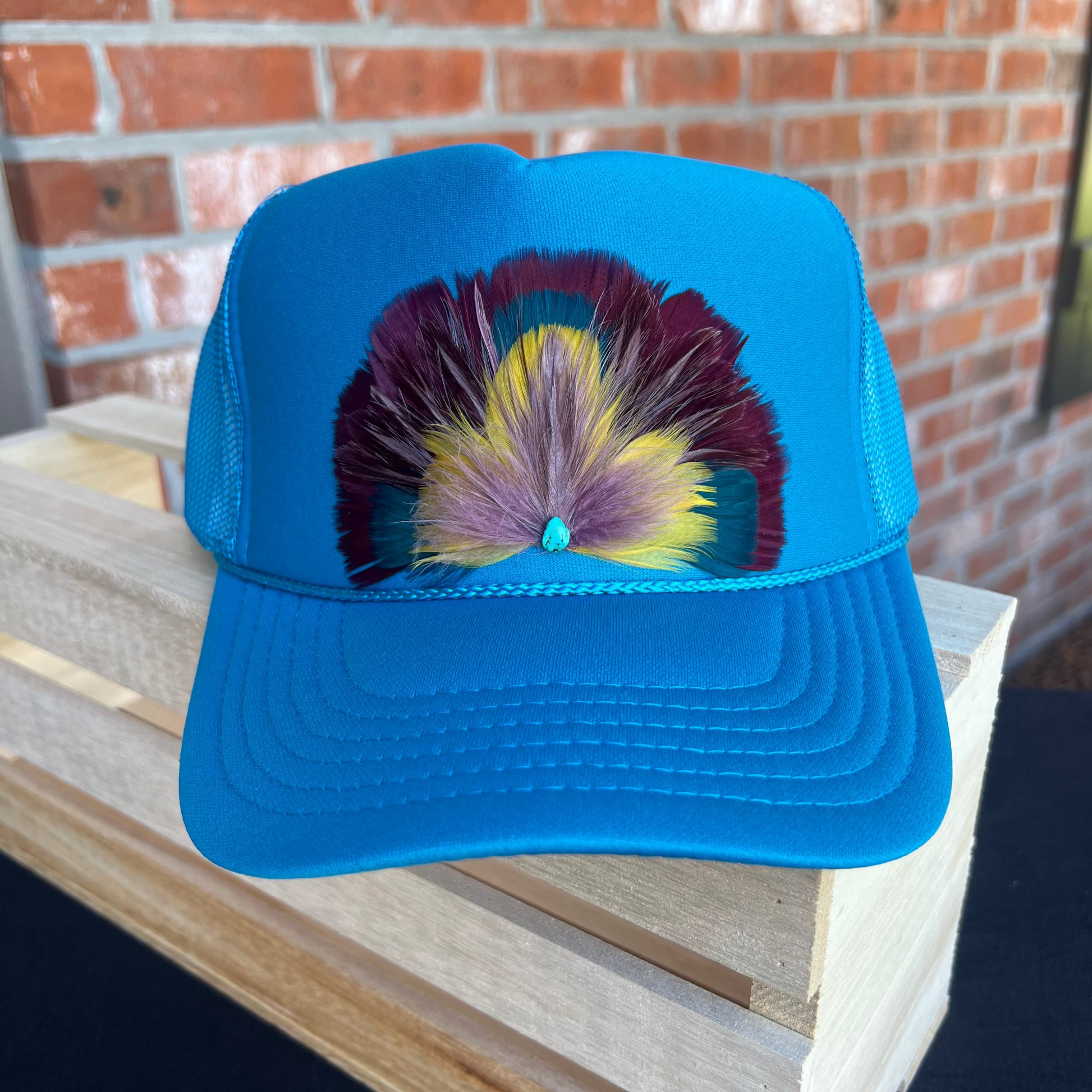 Blue cap with colorful feather design on a wooden surface with a brick wall background