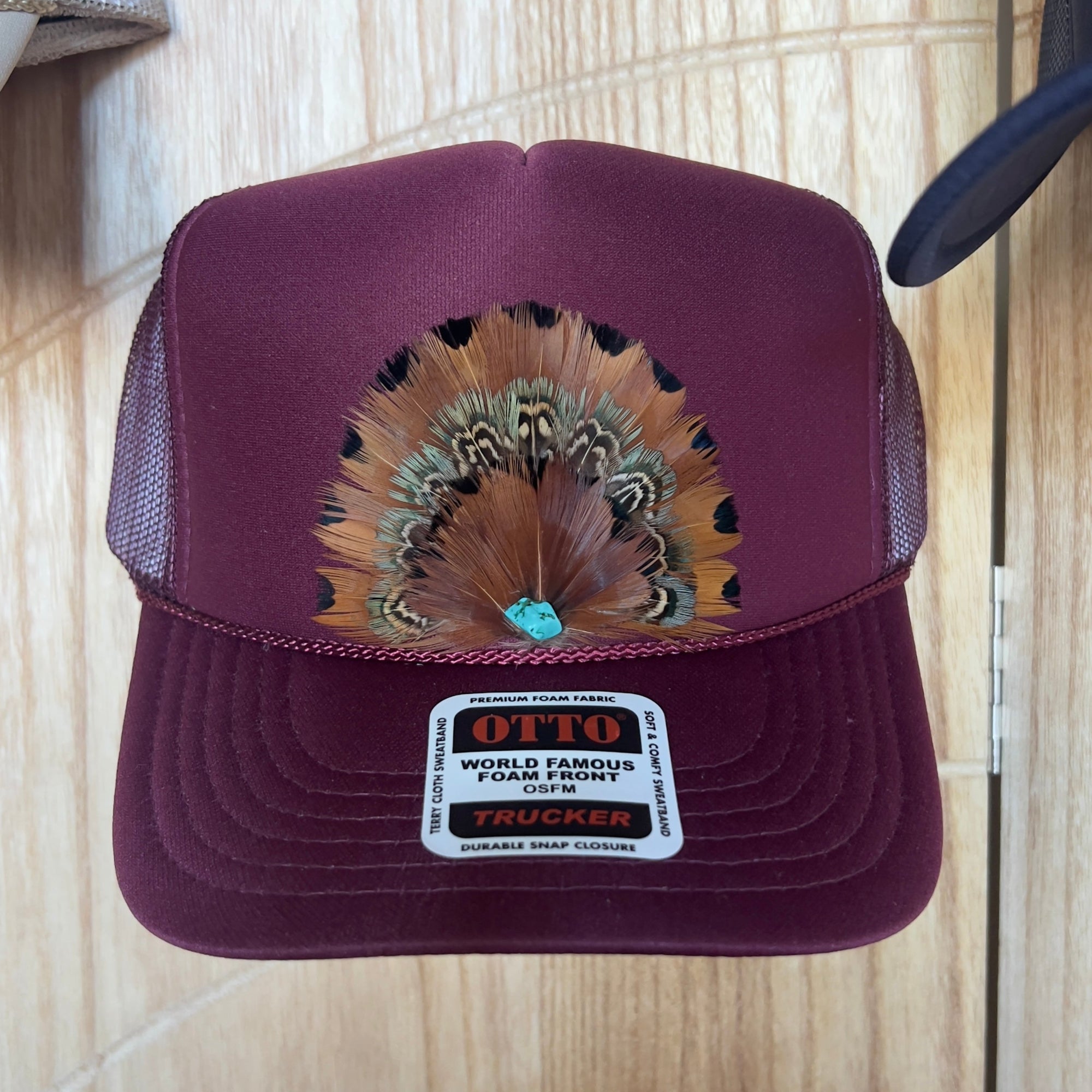 Maroon trucker hat with feather design and Otto label on a wooden surface