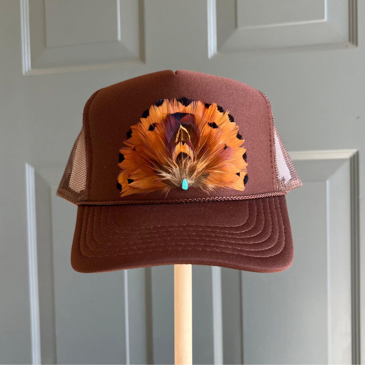 Brown cap with feather design on a stand against a gray wall by Volant Style in Lafayette, LA
