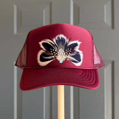 A burgundy trucker hat with a feather design on the front, displayed on a wooden stand against a gray paneled door.