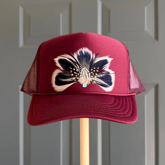 A burgundy trucker hat with a feather design on the front, displayed on a wooden stand against a gray paneled door.