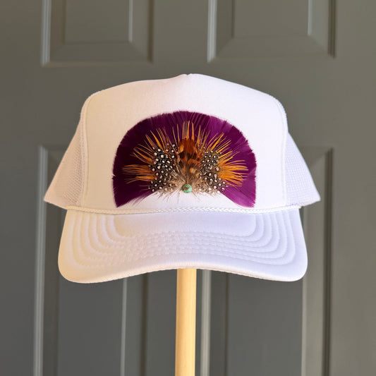 A white trucker-style cap displayed on a wooden stand in front of a dark gray paneled door. The front of the cap features a purple and orange feather fan embellishment.