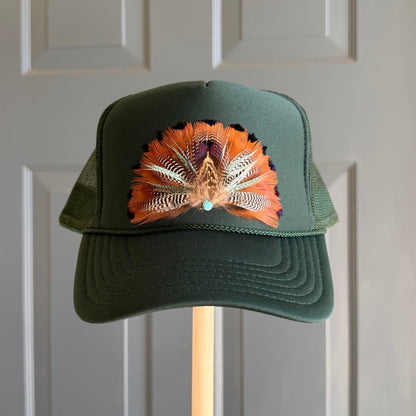 A dark green trucker hat with a colorful feathered design on the front, mounted on a wooden stand in front of a gray paneled door.