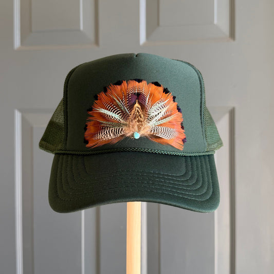 A dark green trucker hat with a colorful feathered design on the front, mounted on a wooden stand in front of a gray paneled door.