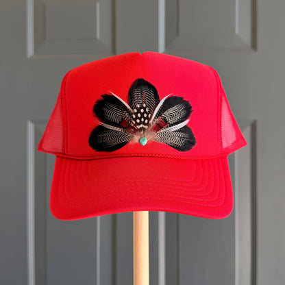 A bright red trucker hat with a black and white feathered floral design on the front, displayed on a wooden stand against a gray paneled door.