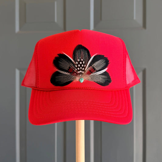 A bright red trucker hat with a black and white feathered floral design on the front, displayed on a wooden stand against a gray paneled door.